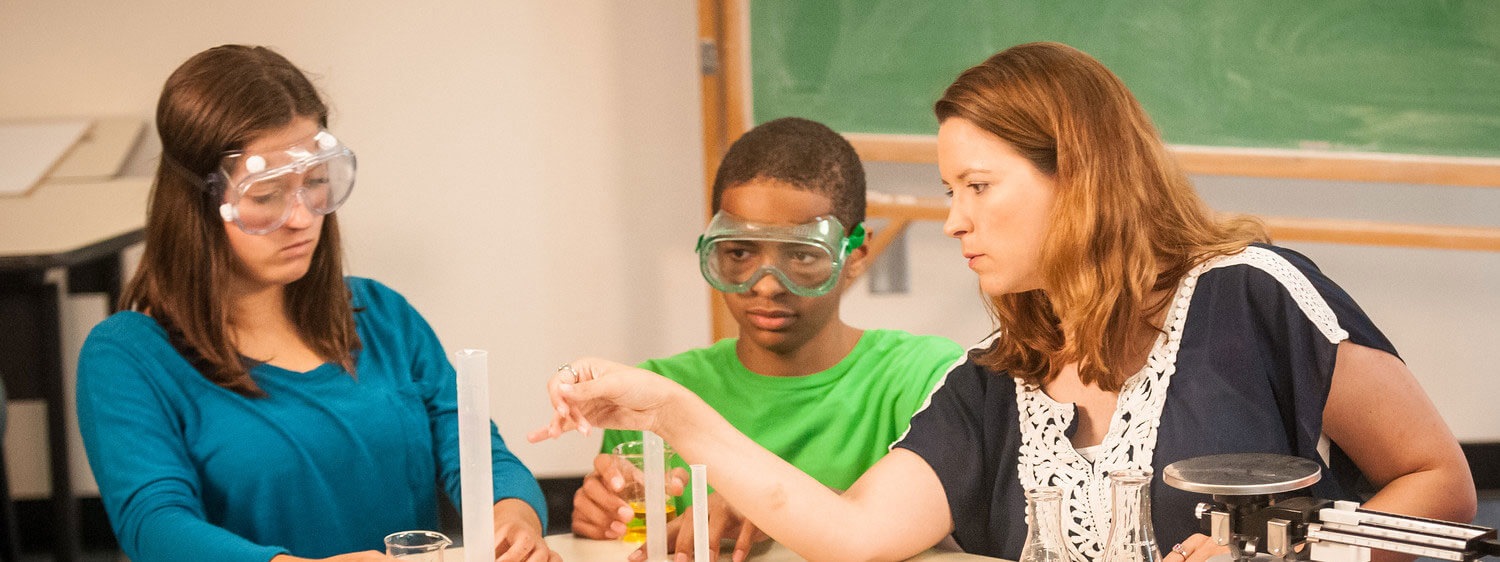 A teacher instructs two students on a science experiment.