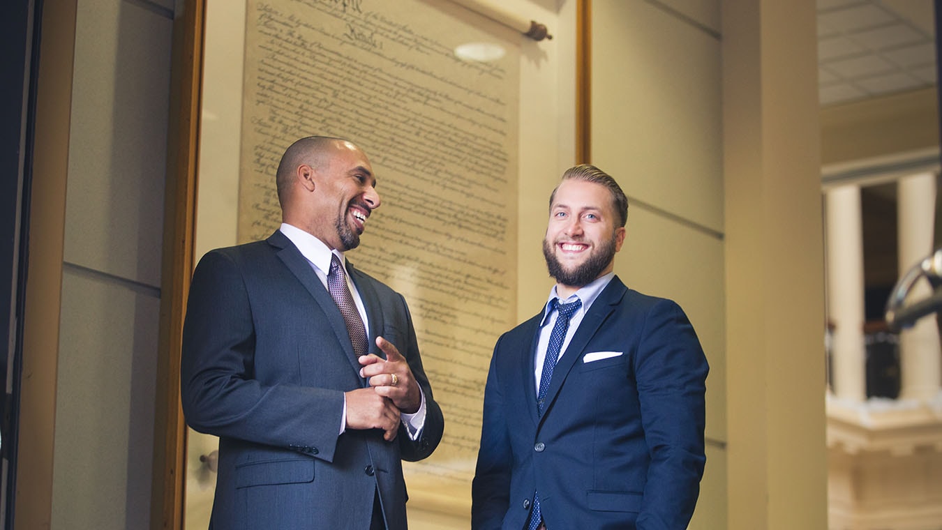 Two students dressed in suits laugh inside Regent University's Robertson Hall, which houses the Government and Criminal Justice degree programs.