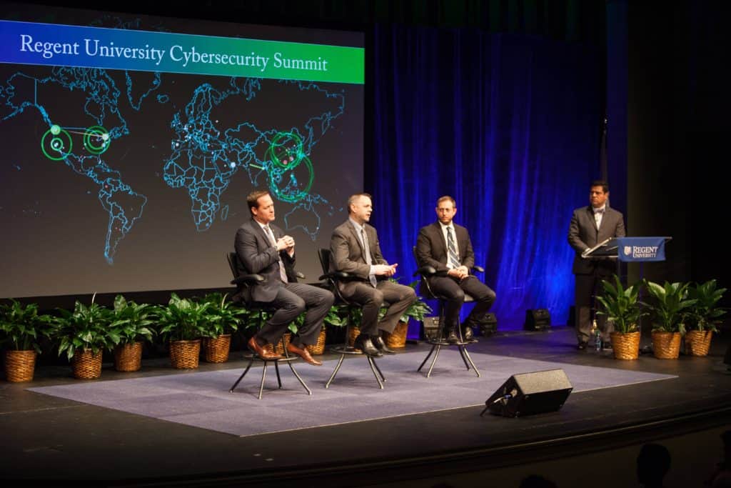 Panelists discuss cybersecurity on stage at Regent University beneath a screen showing a global map with digital attack zones.