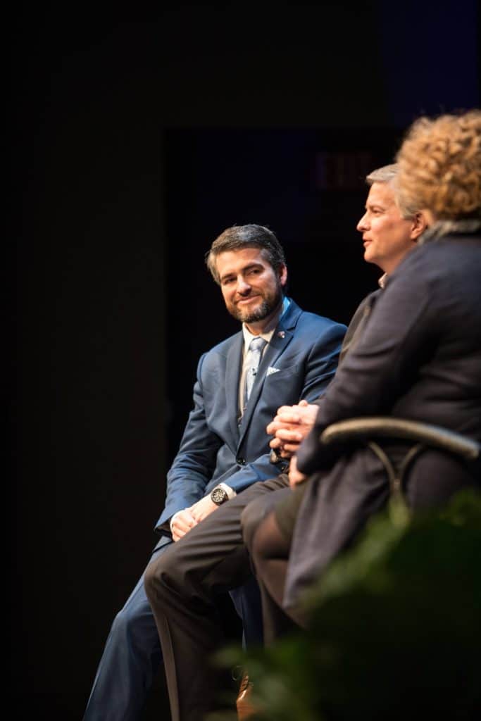 Panelist in a navy suit smiles during a discussion at Regent’s Cybersecurity Summit, seated next to fellow speakers.