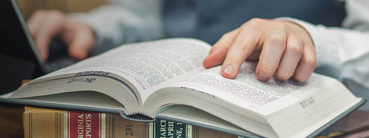 An student's finger on an open book: Pursue an MDiv Biblical Languages degree online or on campus at Regent University, VA 23464.