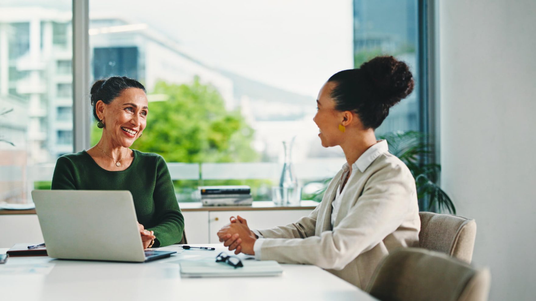 Two professional women having a positive conversation in a modern office setting, one seated behind a laptop in a green sweater, the other in a light blazer, with large windows and greenery in the background