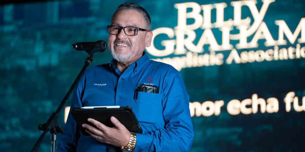 Mario Samaniego, a chaplain and Master of Divinity student at Regent University, speaking on stage at a Billy Graham Evangelistic Association event. He is wearing a blue shirt labeled 'Chaplain' and holding a tablet.