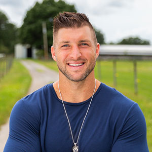 Tim Tebow in standing outside in a blue shirt.