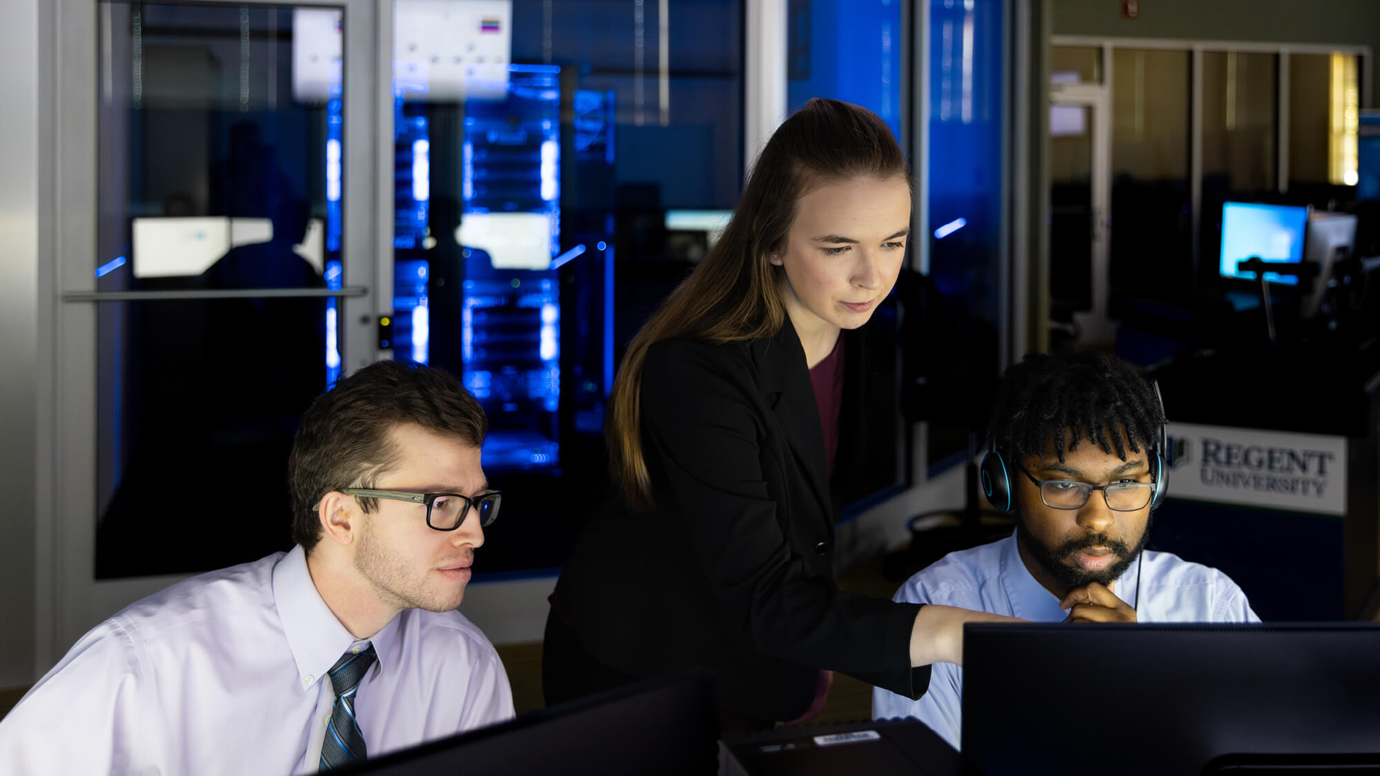 Regent University cybersecurity students collaborating at computer workstations in a dimly lit network operations lab