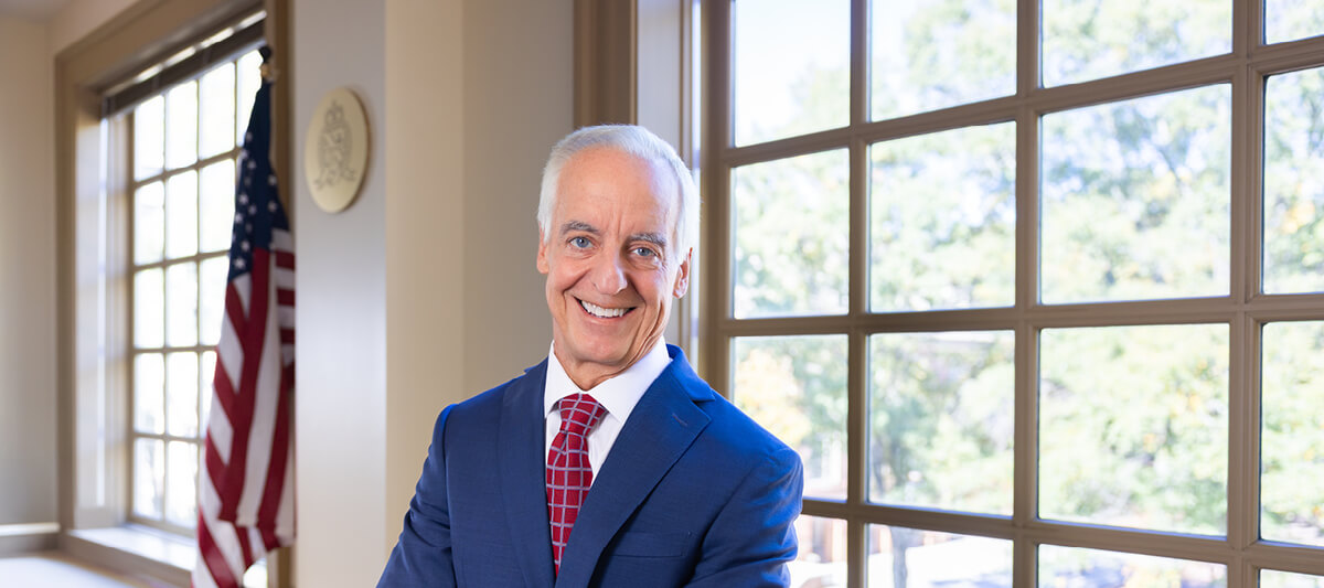 Randy Singer smiling in a professional portrait, wearing a blue suit and red patterned tie, standing in an office with an American flag and large windows in the background
