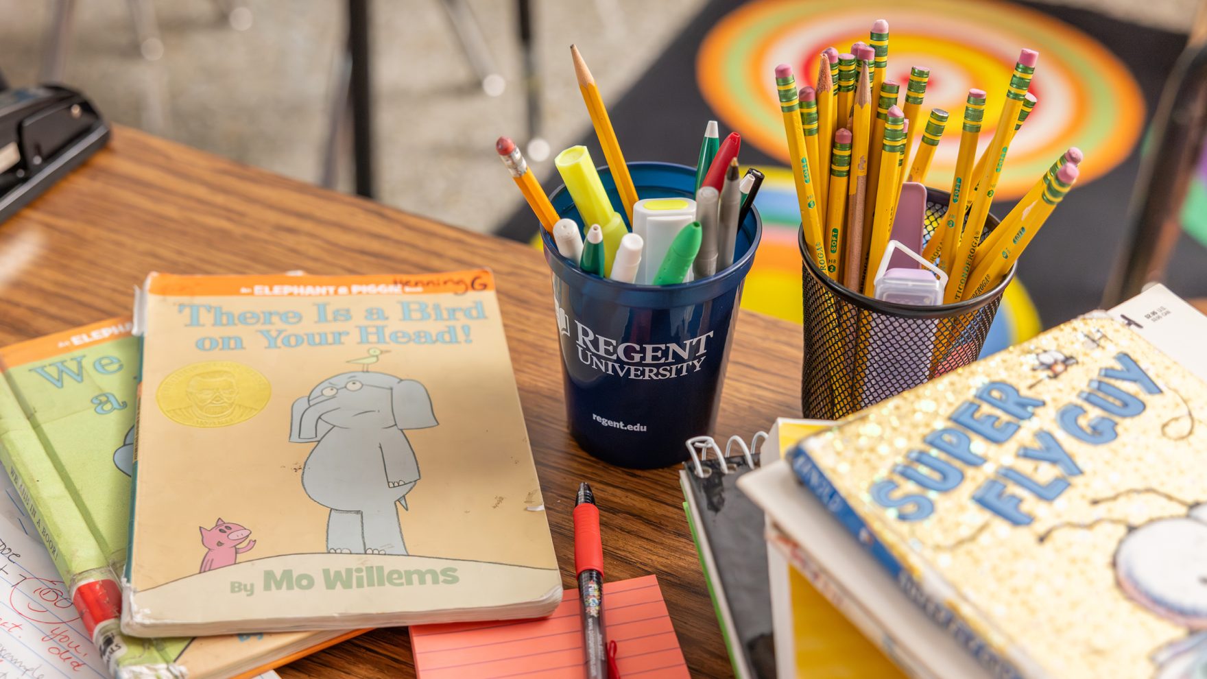 A classroom desk with pencil holders, pencils, highlighters, books, and a notepad on it.
