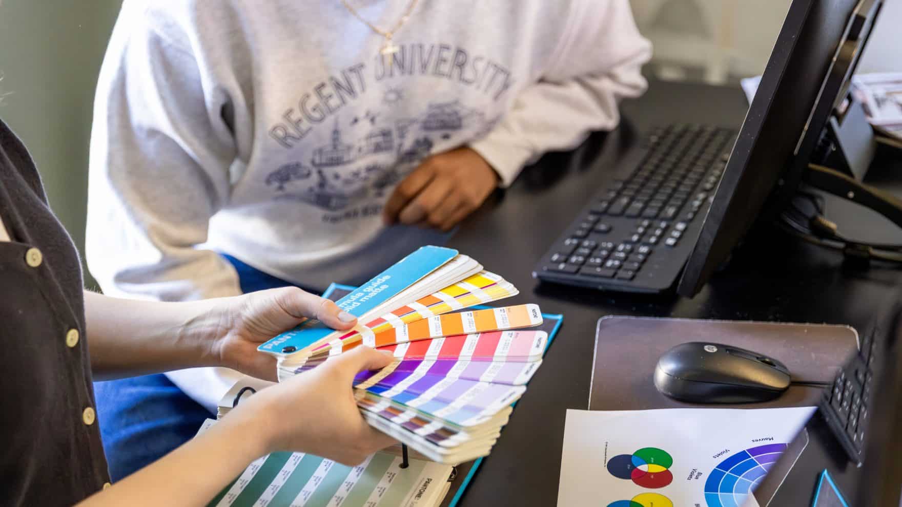 Regent student holding color swatches and Pantone color guide while reviewing graphic design materials at a workstation