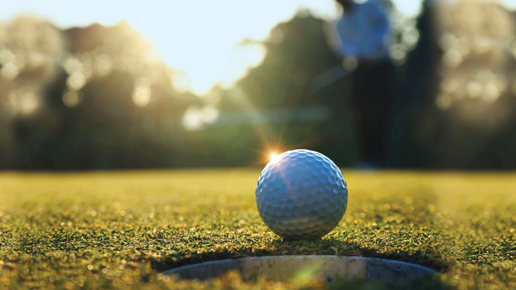 White golf ball sitting next to the golf cup on the putting green of a golf course, as the sun sets in the background.