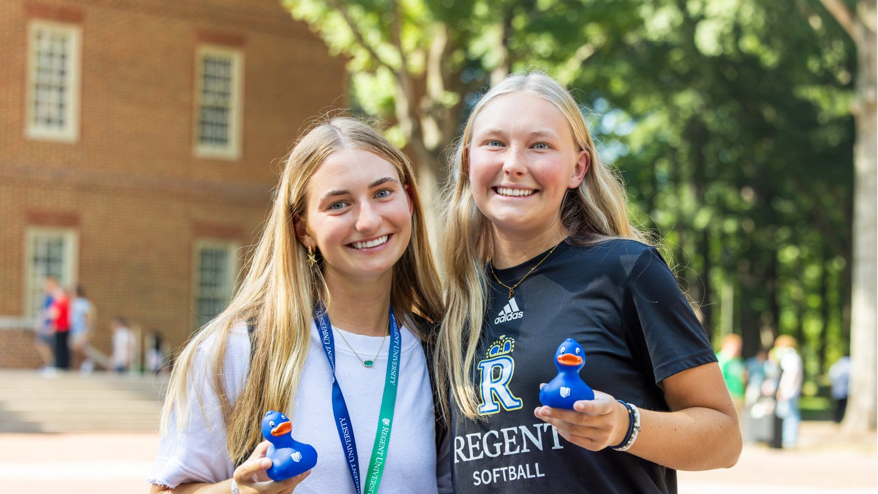 Two students, one wearing a blue and green Regent University lanyard, one wearing a black shirt with Regent Royals logo on it, holding blue rubber ducks while standing outside in front of the Regent University library.