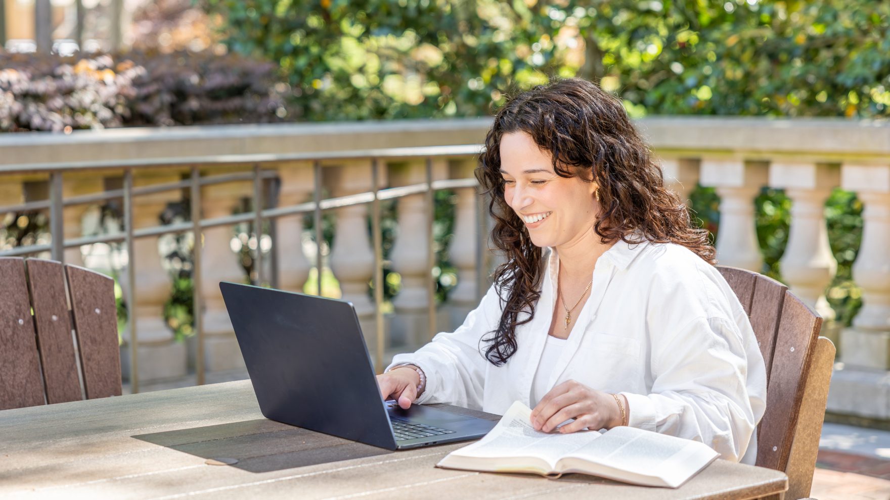 A smiling woman with curly dark hair sits at an outdoor table, typing on a laptop with an open book beside her, with a stone balustrade and green trees in the background.