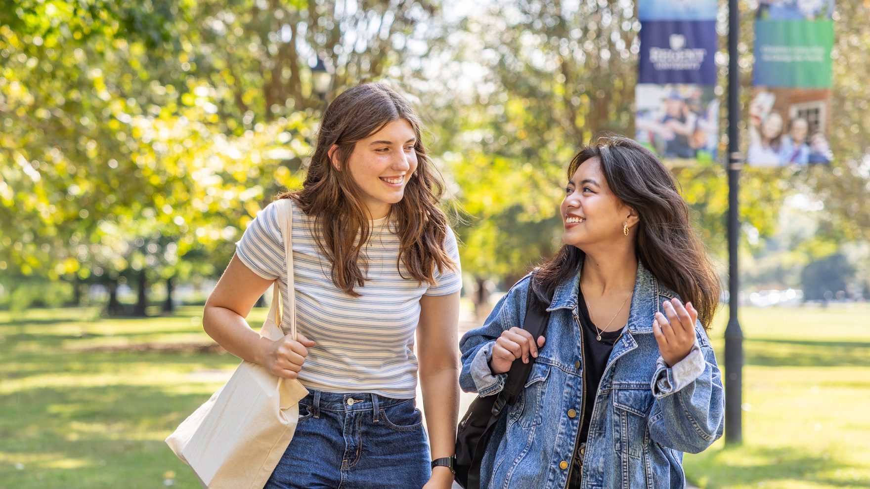 Two smiling female students walk together outdoors on a tree-lined campus lawn, one carrying a tote bag, with a Regent University banner visible in the background.