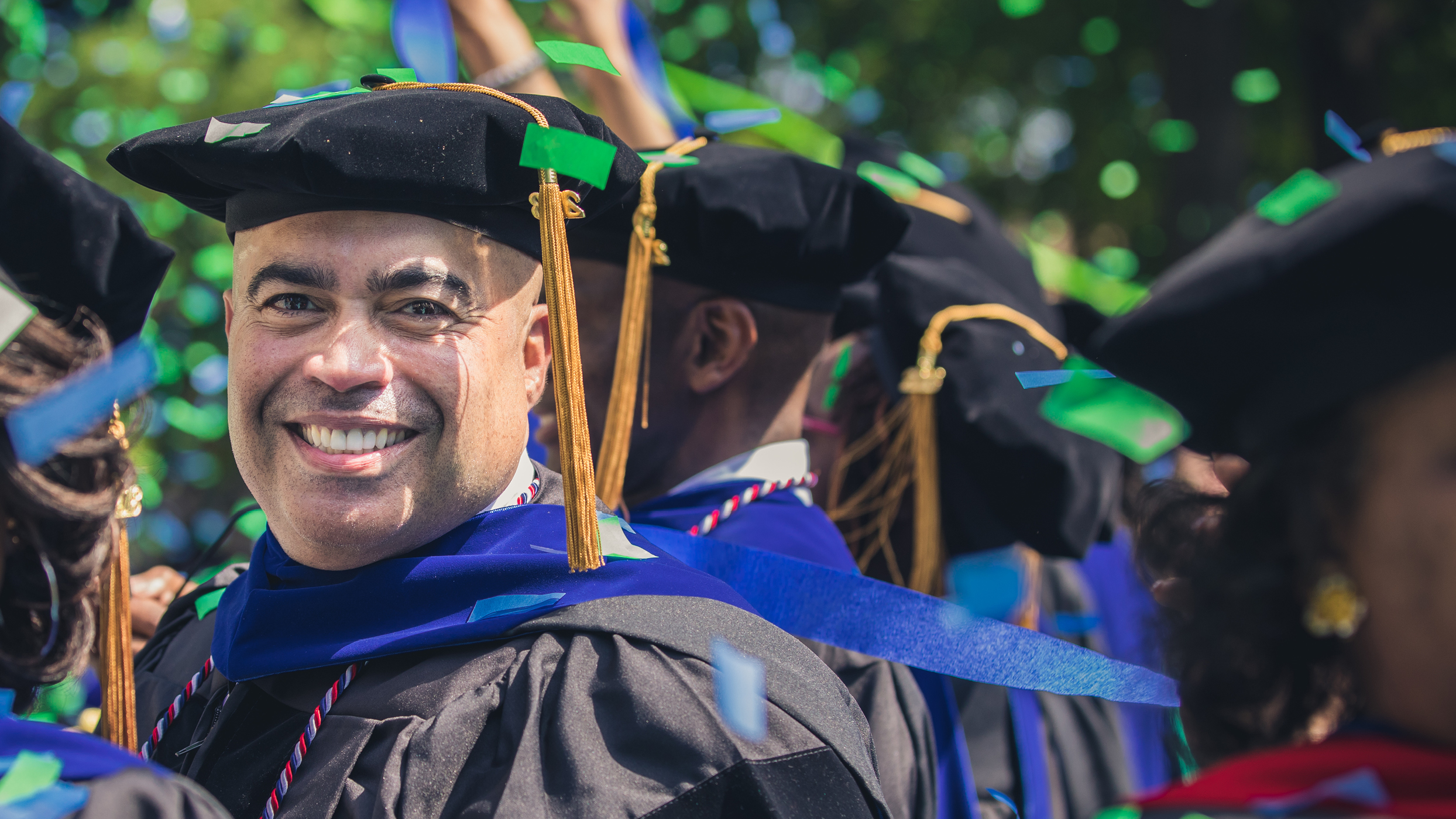 A smiling man in a graduation cap and gown with gold tassels stands among fellow graduates outdoors at a Regent University commencement ceremony.