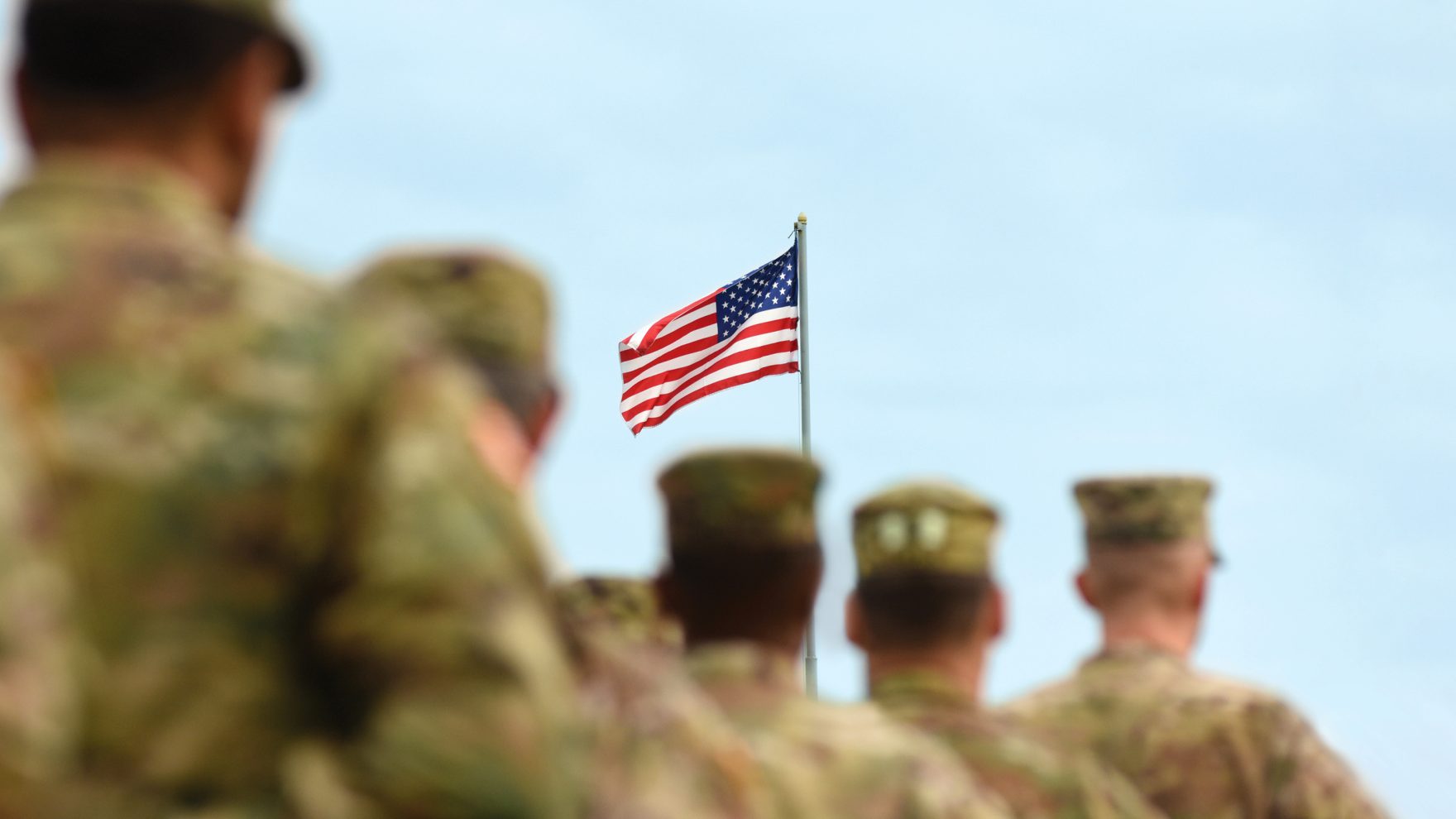 A row of service members in camouflage uniforms and caps stand facing an American flag on a pole against a blue sky.