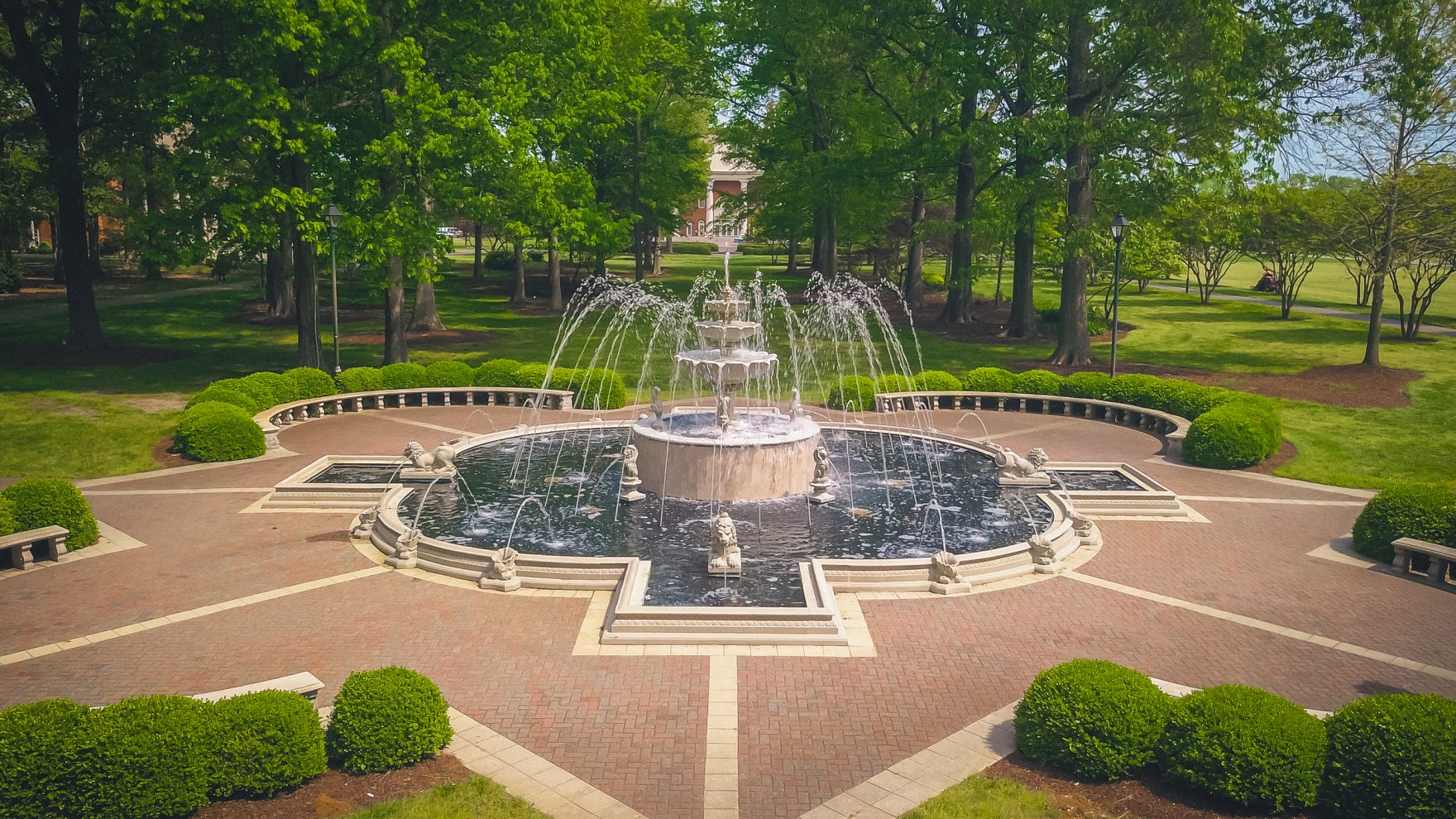 An aerial view of a tiered stone fountain surrounded by manicured hedges and brick pathways on the Regent University campus, with trees and a building in the distance.