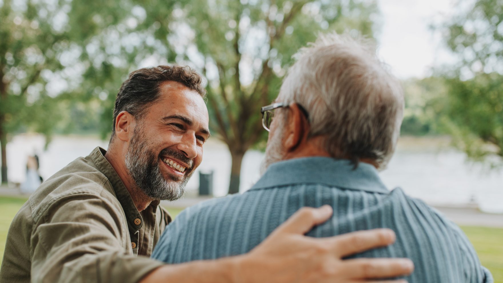 Middle aged man, smiling with his hand on the back of an elderly man, both sitting and talking outside in a park.