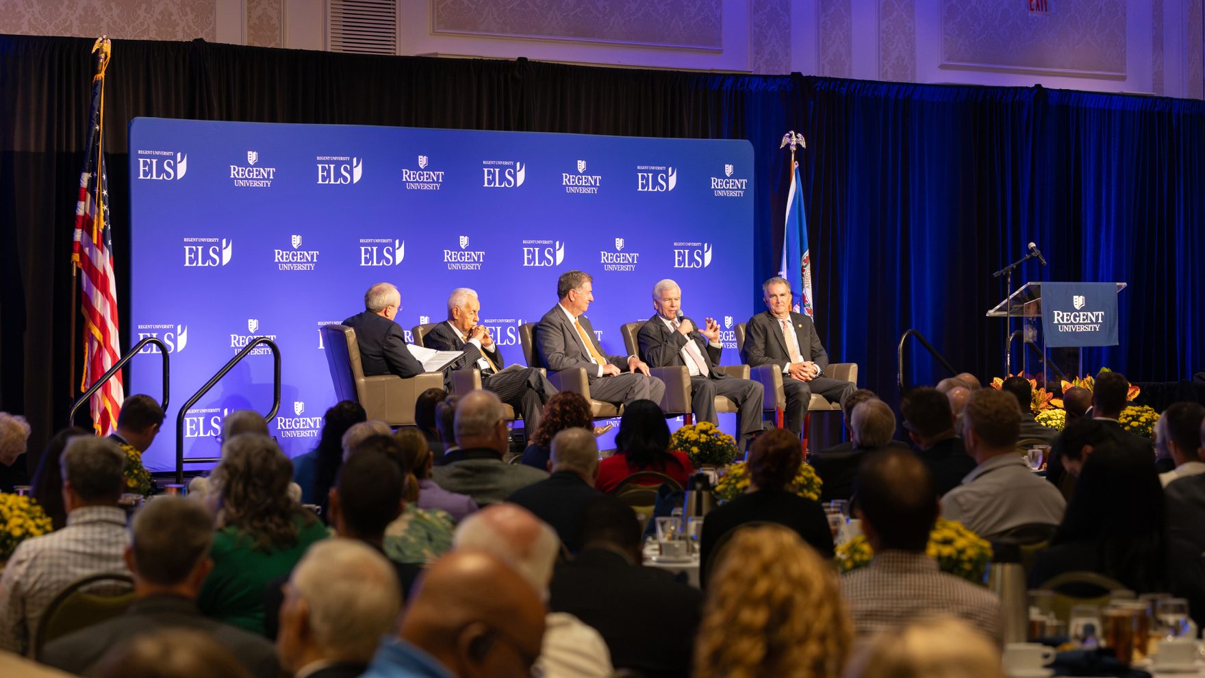 Four former governors of Virginia sitting on a platform in front of a backdrop, facing a large crowd of business professionals as panelists at an Executive Leadership Series event held at Regent University.