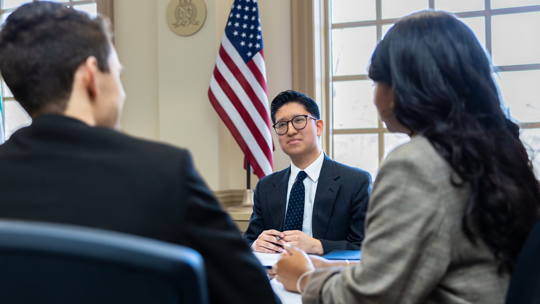 A man in a suit and glasses sits at a table across from two colleagues, with an American flag and a wall seal in the background, suggesting a government or policy meeting.