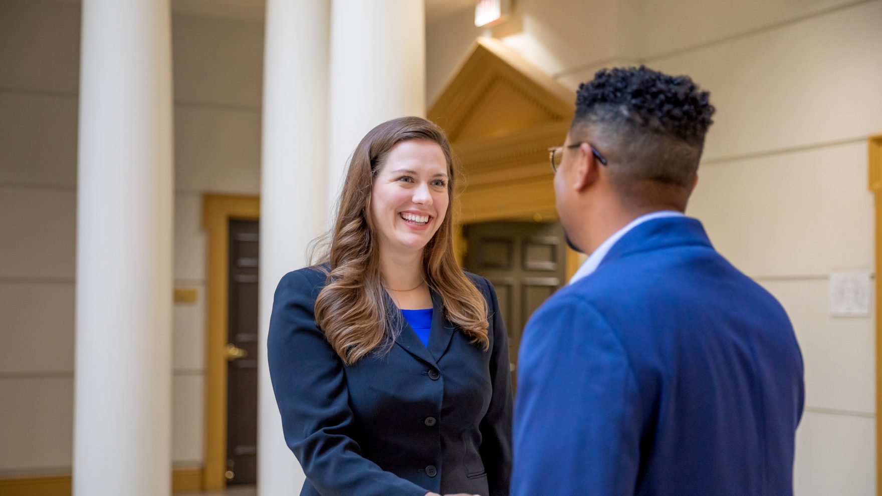 A smiling woman in business attire shakes hands and talks with a man in a blue suit in a bright Regent University hallway with white columns.