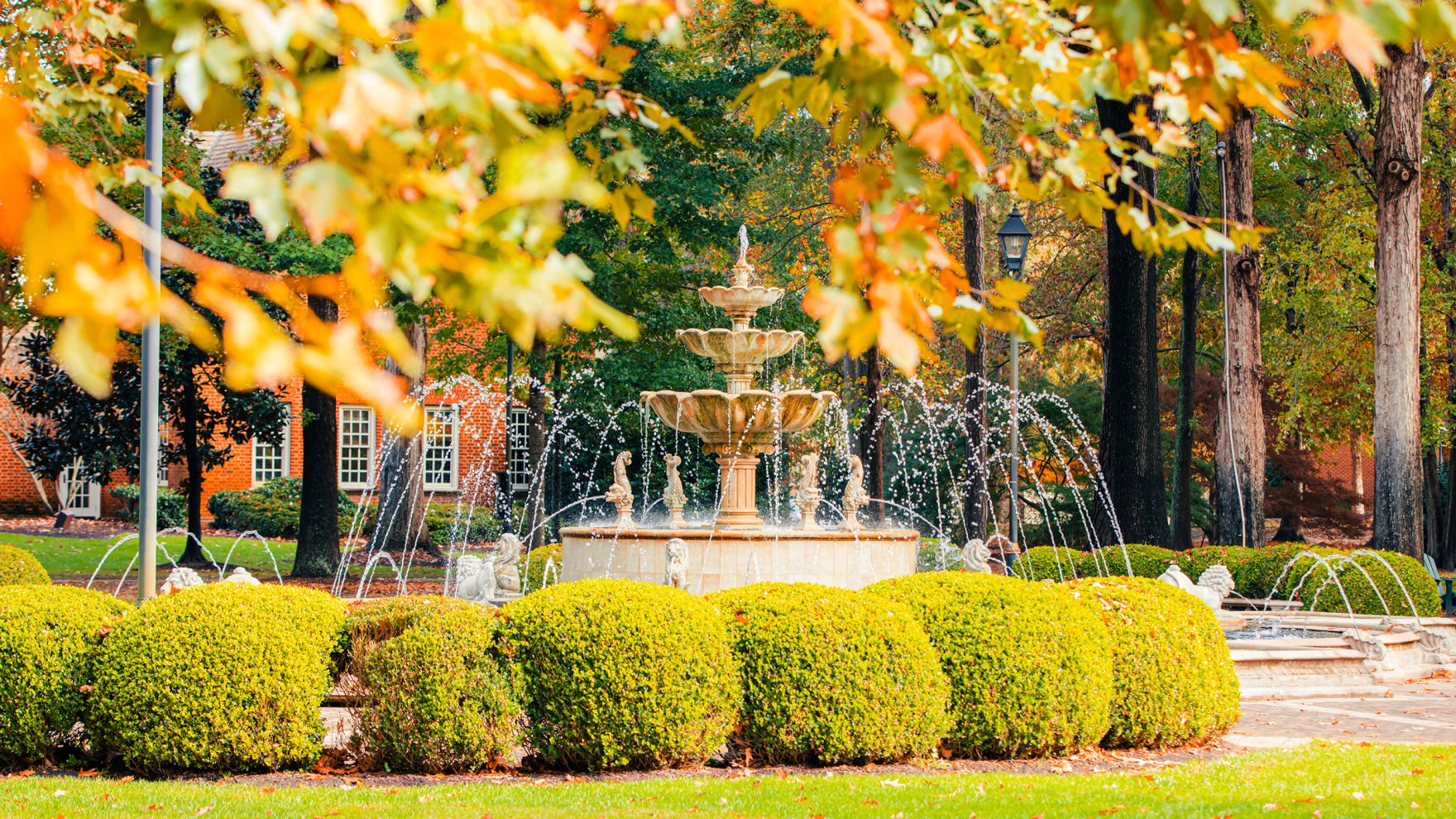 A stone tiered fountain surrounded by rounded green hedges on a campus lawn, framed by golden autumn leaves and a brick building in the background.
