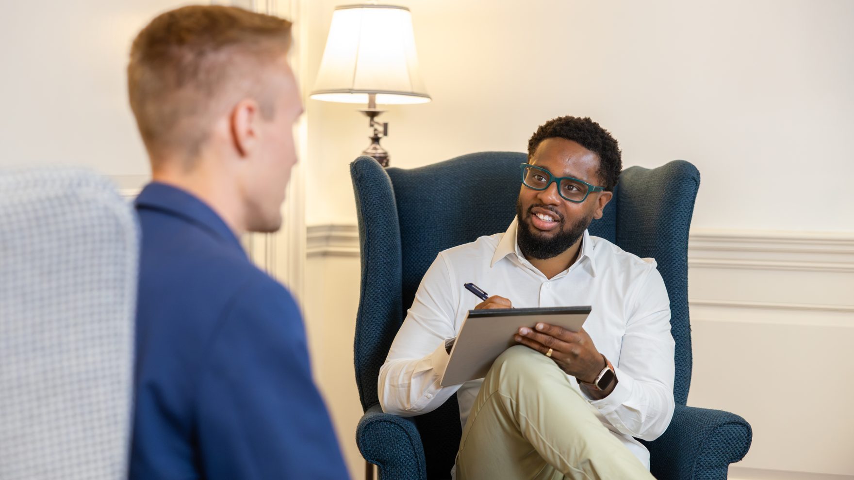 A man with glasses and a beard sits in a wingback chair taking notes on a tablet while talking with another man seated across from him, evoking a counseling session setting.