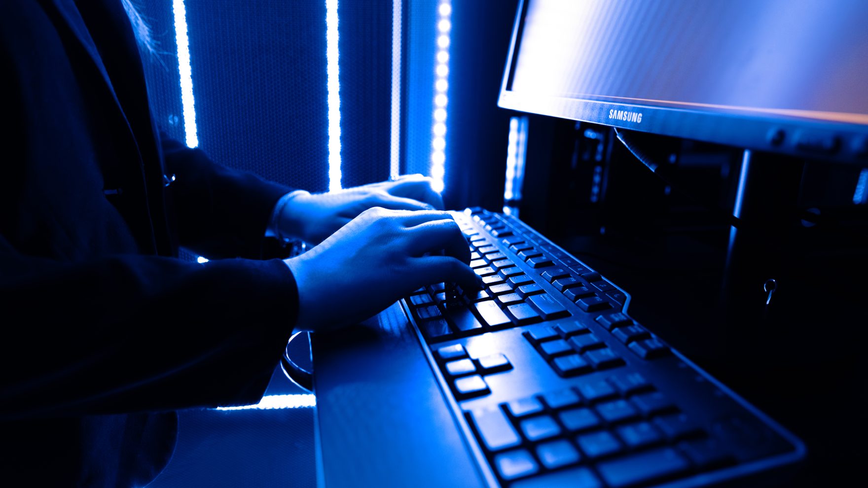 Regent University student sitting in front of a computer, typing on its keyboard while looking at the monitor in a room lit-up by a blue lamp.