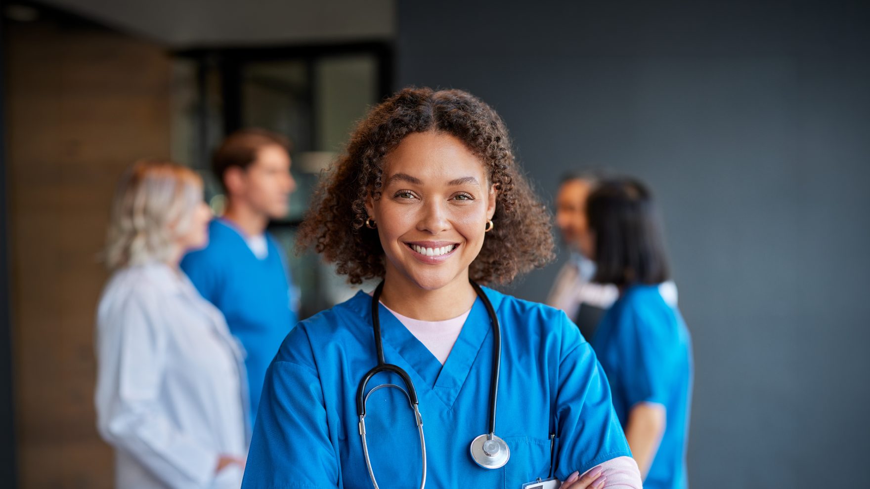 A smiling woman in blue scrubs with a stethoscope around her neck stands with arms crossed in a medical facility hallway, with colleagues in scrubs and a white coat talking in the background.